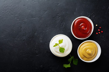 Obraz na płótnie Canvas Set of three sauces - mayonnaise, mustard and ketchup in white ceramic bowls on black stone or concrete background. Selective focus. Top view.