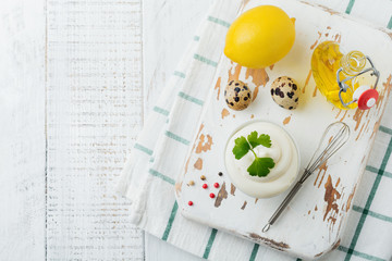 Traditional mayonnaise sauce in white ceramic bowl and ingredients for its preparation on white wooden background. Selective focus.