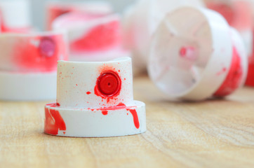 Several plastic nozzles from a paint sprayer that lie on a wooden surface against a gray wall background. The caps are smeared in red paint. The concept of street art and graffiti © mehaniq41