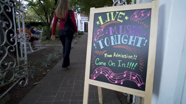 Beautiful blonde woman in yoga pants walks into a garden with a sign for live music.