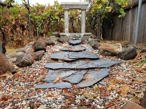 A stone water fountian with classic style pillars granet plates stream into ;lava and river rock with a green vineyard boardering a brown wooden fence