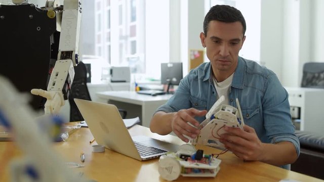 Smart worker. Experienced constructor creating robot parts while sitting at the table with a laptop and being attentive