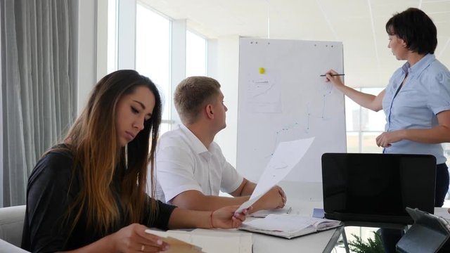 mentor near chart of business development on Whiteboard speaks with young partners in office with large panoramic windows