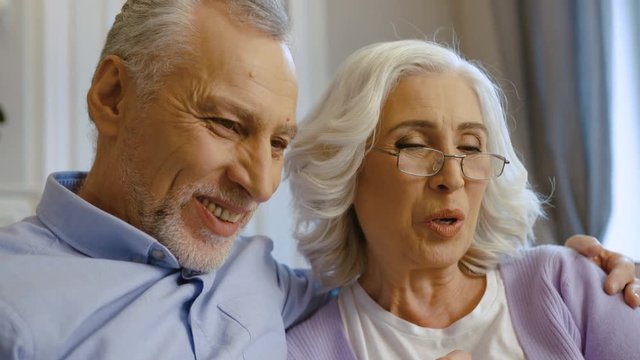 Close up shot of old couple sitting on the sofa at home and possing on camera when making selfie.