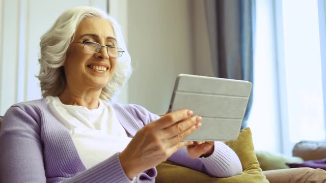 Close up shot of beatiful old woman in glasses using tablet for surfing in the internet at home in the living room.