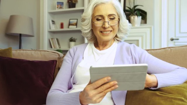 Close up shot of smiling old woman in glasses using tablet while sitting on sofa at home in the living room.