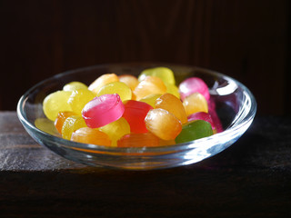Small multi-colored sweets on a glass saucer on a dark wooden background. Dark light.