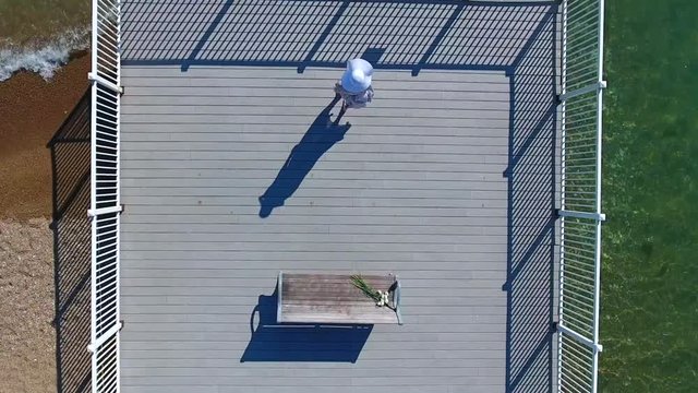 Woman is standing up from the bench and walking on the wooden pier to the fence. Woman is wearing white sun hat on the pier. Shooting from above.