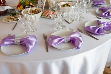 Served table plate with purple cloth. Wedding tables set for fine dining or another catered event. Napkin in the form of a bow. Plate, fork, knife on the background of the snacks on the table.