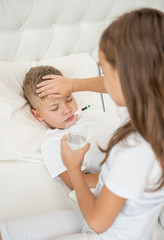 A girl with a glass of water touches her brother's forehead, checking the temperature