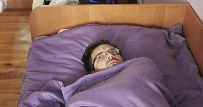 A sick young man with glasses lies in bed and measures the temperature.