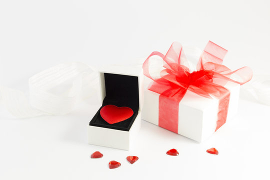 A red velvet heart in a white jewelry box, a white gift box with red ribbon and small shiny red decorative hearts on white background.