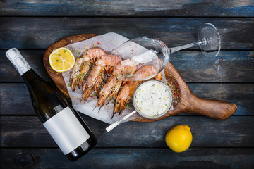 Large shrimp or langoustine with white sauce, bottle of wine, glass for the wine and half a lemon on a wooden board. Top view