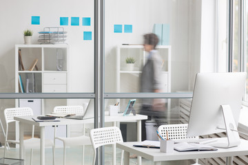 Blurred human figure moving behind transparent wall between two offices and empty workplace on foreground