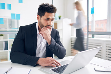 Pensive man looking at laptop display while brainstorming and solving problems of company