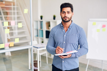 Fototapeta premium Young confident office worker with pen and notebook looking at camera on background of his workplace and whiteboard