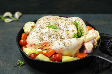 Whole raw chicken in black frying pan placed with fresh vegetables and herbs on dark background