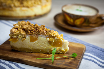 Slice of cheesecake with peach on the wooden cutting board. Cake and cup of coffee on the background