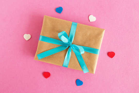 Gift box wrapped of craft paper and blue ribbon with colorful hearts on the pink background, top view.
