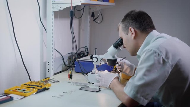 A man who looks through a microscope holds a pencil in his hand, he wore gloves to avoid leaving traces on the microcircuits