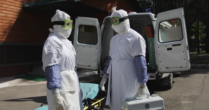 Two unrecognizable doctors or nurses wearing full Ebola virus protection uniform  standing near ambulance car and talking.