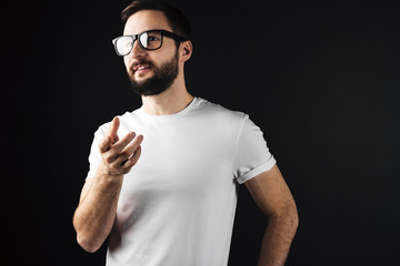 Naklejka premium Handsome bearded young hipster wearing glasses and white bkank tshirt and shows something hand
