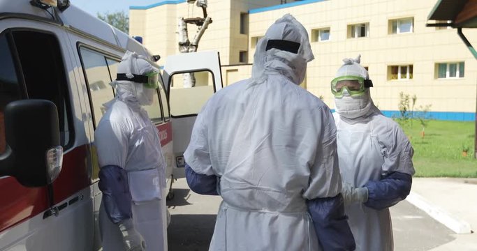 Three unrecognizable doctors or nurses wearing full Ebola virus protection uniform &nbsp;standing near ambulance car and talking.