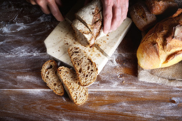 Hands cutting rye bread on a wooden brown board. Integral bread with sunflower, sesame, flax and poppy seeds. Top view