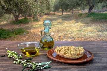 Bowl and bottle with extra virgin olive oil, olives, a fresh branch of olive tree and cretan rusk dakos on wooden table, in an olive tree field at Crete, Greece. - obrazy, fototapety, plakaty
