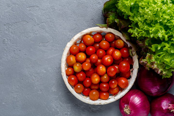 Fresh healthy diet colorful ingredients for a salad, bowl of cherry tomatoes with green salad lettuce and red onions on grey concrete background table. Top view with copy space.

