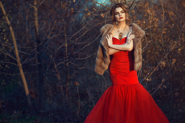 Fototapeta premium Gorgeous glam model with updo hair and beautiful make up wearing posh red fishtail dress and luxurious mink vest standing in dry autumn bushes with crossed arms. Vintage necklace on her neck