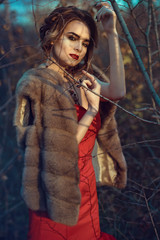 Gorgeous glam model with updo hair and beautiful make up wearing red dress standing in dry autumn trees at dawn. Luxurious mink vest slipped over her shoulders. Shadows of branches on her face