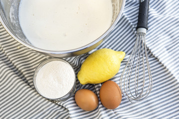 cuenco con leche para hacer uncake con los ingredientes de limón,huevos,harina y un batidor en un mantel de rayas azules y blancas.