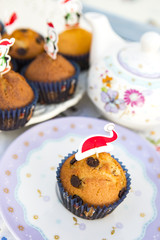plato decorado con una magdalena con trozos de chocolate y un gorrino rojo de navidad como adorno y en el fondo otro plato de magdalenas y una tetera difuminadas