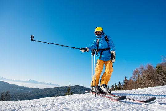 Full length shot of a skier standing on top of a mountain on a beautiful sunny winter day taking a selfie with action camera on monopod. Blue sky and winter forest on the background
