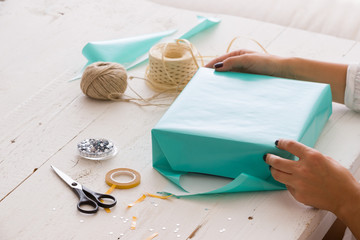 Closeup of woman's hands with gift ribbon, twine, tape and present wrapped in turquoise gift paper on white wooden background. Christmas, birthday or any other celebration preparations.