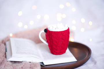 Knited cup of coffee on open book over Christmas lights close up. Good morning.