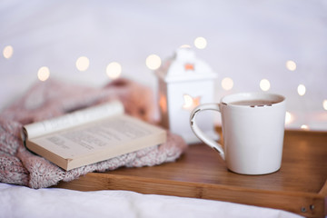 Mug of coffee with opn book and knitted sweater on wooden tray close up over Christmas lights. Good...