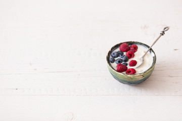 Top view on small bowl with organic yogurt with blueberries and raspberries on white wooden background. Girl having healthy breakfast at home. Healthy eating, snack.