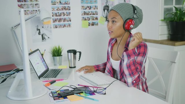 Young ethnic woman wearing hat and listening to music with headphones posing at table in office working on new project and watching computer.