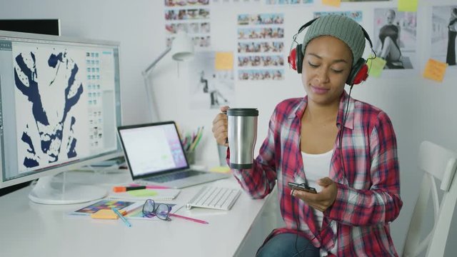 Young ethnic hipster woman in headphones enjoying cup of coffee and relaxing with smartphone while sitting at table in office.