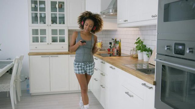 Wonderful causal woman listening to music with headphones and smartphone and dancing happily holding glass of fresh juice.