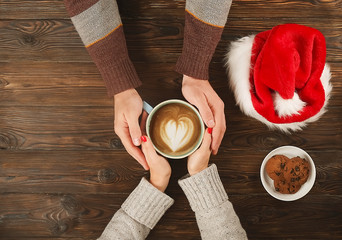 Lovely couple holding cup of coffee in hands on wooden background with santa hat and chocolate cookies top view