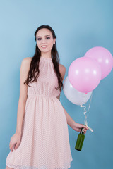 A young girl in a flower dress standing surrounded by balloons and held a green bottle