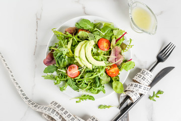 Healthy balanced diet concept, weight loss, calorie counting. Plate with green salad leaves, tomatoes, avocado with yogurt dressing, white table, with fork, knife, measuring tape, top view copy space
