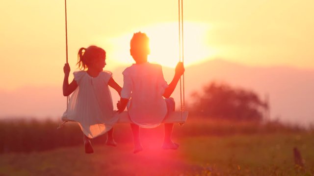SLOW MOTION SILHOUETTE, LENS FLARE: Couple kids holding hands while swinging on swing in gold evening. Unrecognizable kids swaying on late afternoon. Young boy and girl in love holding hands on swing.