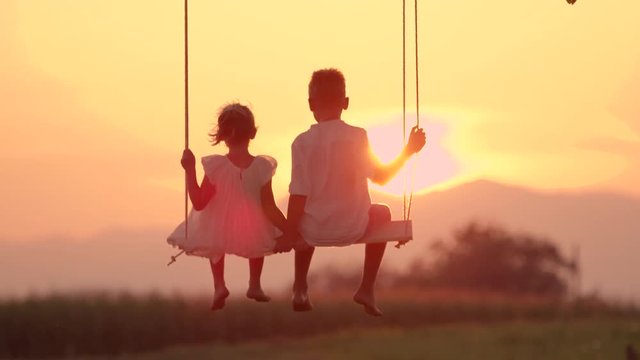 SLOW MOTION SILHOUETTE, LENS FLARE Two kids looking at sunset while swinging on swing in gold evening. Unrecognizable kids swaying on late afternoon. Little brother and sister enjoying gold sundown.