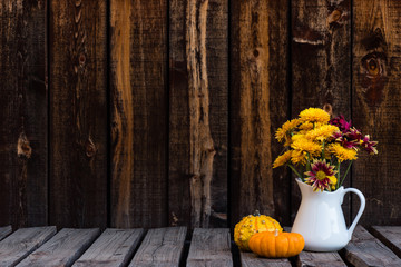 White pitcher holding a bouquet of fall colored mums and two gourds on a rustic wooden table.