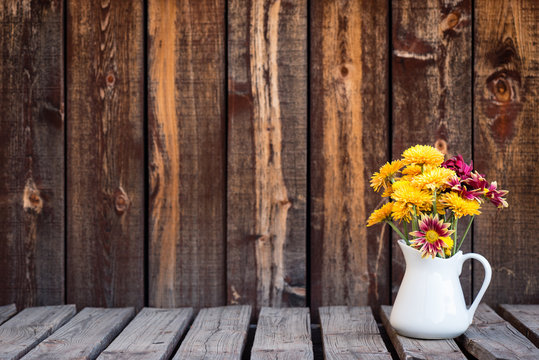 White pitcher holding a bouquet of fall colored mums on a rustic wooden table.