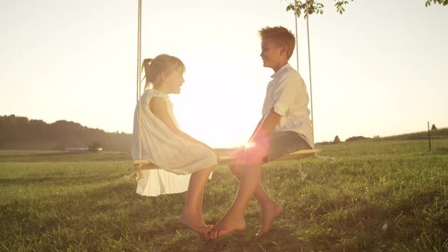 SLOW MOTION CLOSE UP Smiling brother and sister sitting on wooden swing having fun at sunset. Couple of playful kids facing each other talking on swing. Cheerful boy and girl enjoying summer evening.